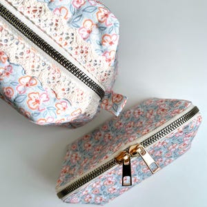 May include: Two floral print zippered pouches with white lace trim. The larger pouch is closed and the smaller pouch is open, showing the gold zipper pull.