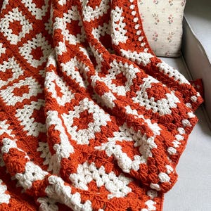 May include: A handmade crochet blanket in a geometric pattern of white and orange squares. The blanket is draped over a gray sofa, with a floral pillow in the background. The blanket is made of soft yarn.
