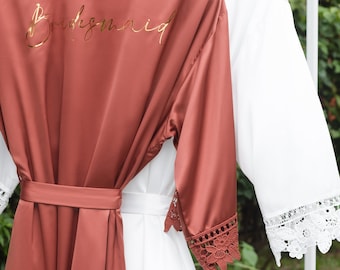 May include: Two satin robes with lace trim. One robe is rust colored and has the word "Bridemaid" embroidered on the back. The other robe is white.