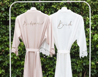 May include: Two white satin robes with lace trim, hanging on a white rack. One robe says "Bride" in gold script, the other says "Bridesmaid" in gold script.