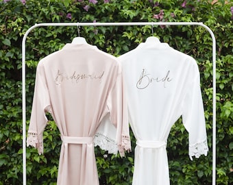 May include: Two white satin robes with lace trim hanging on a white rack. The robe on the left is blush pink and says "Bridesmaid" in gold script. The robe on the right is white and says "Bride" in gold script.