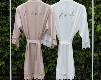 May include: Two satin robes hanging on a white rack. The robe on the left is blush pink with the word "bridesmaid" embroidered on the back. The robe on the right is white with the word "bride" embroidered on the back. Both robes have lace trim.