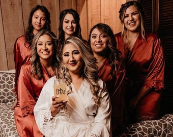 May include: A group of women wearing matching robes, with one woman in the center wearing a white robe and holding a glass with the word "Mrs." on it. The women are sitting on a bed with a gray and white patterned bedspread.