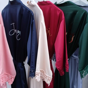 Pu&ograve; includere: Sei accappatoi di raso in diversi colori con bordino in pizzo e ricamo personalizzato. Gli accappatoi sono appesi a un appendiabiti bianco. Il ricamo include i nomi "Joy", "Erin" e "Bridesmaid".