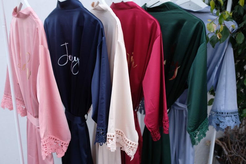 Pu&ograve; includere: Sei accappatoi di raso in diversi colori con bordino in pizzo e ricamo personalizzato. Gli accappatoi sono appesi a un appendiabiti bianco. Il ricamo include i nomi "Joy", "Erin" e "Bridesmaid".