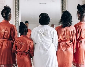 May include: Five people wearing matching robes with gold lettering. The bride is wearing a white robe with "Bride" written on the back. The other four robes are rust colored with the names "Brittany", "Maid of Honor", "Bridesmaid", and "Junior Bridesmaid" written on the back.