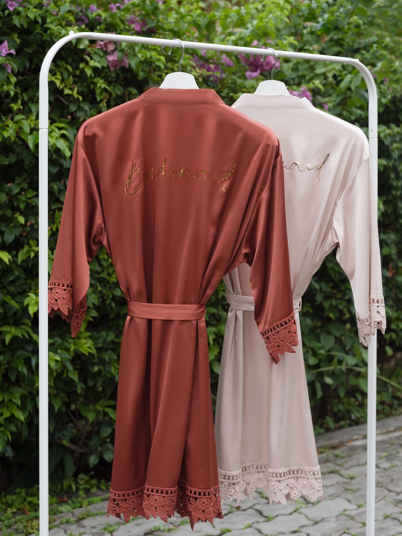 Pu&ograve; includere: Due accappatoi di raso con bordino in pizzo, uno color ruggine con "Bridemaid" ricamato in oro, l'altro color cipria con "Maid" ricamato in oro.