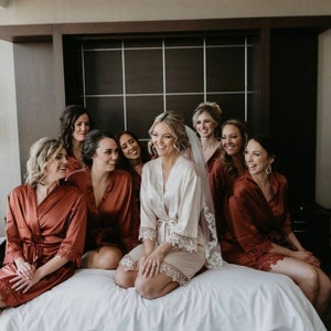 Pu&ograve; includere: Un gruppo di damigelle d'onore che indossano accappatoi di raso color ruggine con bordi in pizzo posano per una foto con la sposa che indossa un accappatoio di raso bianco con bordi in pizzo. La sposa indossa un velo e sorride alla telecamera.
