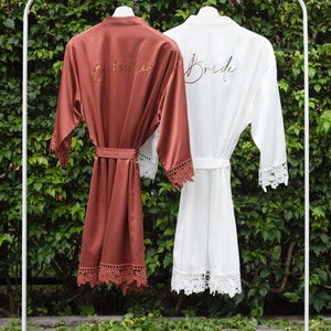 May include: Two satin robes hanging on a white rack. The robe on the left is rust colored with the word "Bridesmaid" embroidered in gold. The robe on the right is white with the word "Bride" embroidered in gold. Both robes have lace trim.