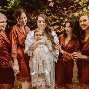May include: A group of women wearing rust-colored satin robes with lace trim. One woman is holding a baby girl wearing a white lace dress. The women are standing in a forest setting.