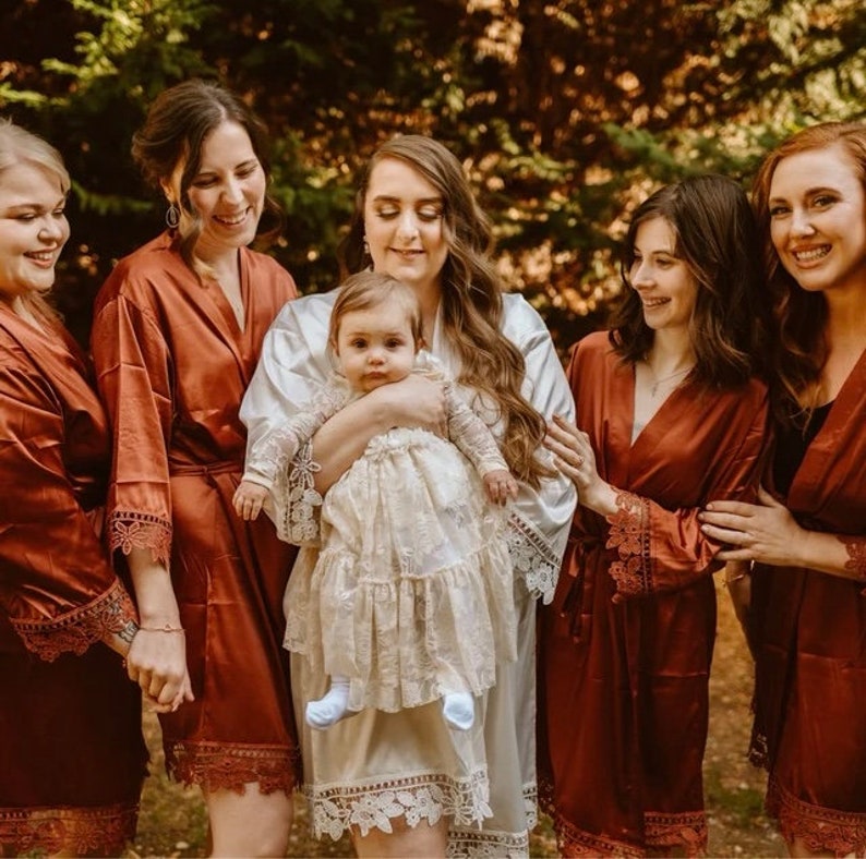 May include: A group of women wearing rust-colored satin robes with lace trim. One woman is holding a baby girl wearing a white lace dress. The women are standing in a forest setting.