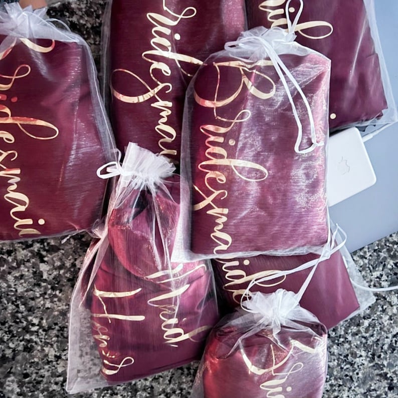 May include: Burgundy satin robes with gold lettering that says "Bridesmaid" in a cursive font. The robes are packaged in clear organza bags with white drawstrings.