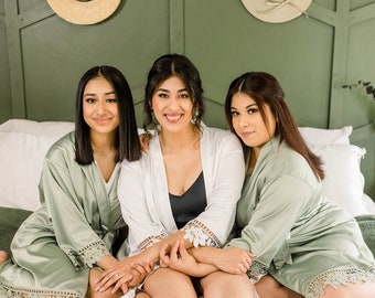 May include: Three women wearing matching sage green satin robes with lace trim. The robes are tied at the waist and have long sleeves. The women are sitting on a white bed with a green wall behind them.