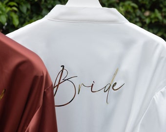 May include: White satin robe with the word "Bride" written in gold script on the back. A burgundy satin robe is partially visible in the background.