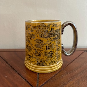 Könnte beinhalten: Gelbe Keramiktasse mit einer schwarzen Karte von East Berkshire, England. Die Tasse hat einen silbernen Henkel und einen geriffelten Boden. Die Tasse steht auf einer dunkelbraunen Holzoberfläche.