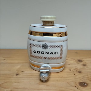May include: White ceramic barrel-shaped decanter with gold trim and a cork stopper. The word "COGNAC" is displayed in the center, framed by a decorative border. A silver spigot is located at the bottom.