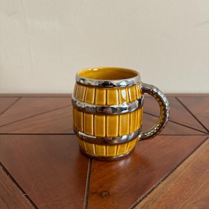 May include: A ceramic mug shaped like a barrel, with a yellow exterior and a silver-colored rim and handle. The mug has a textured surface mimicking wooden staves. The handle is also silver-colored and has a textured design.