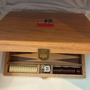 Op de afbeelding: Een houten backgammonset met een lichtbruine afwerking. De set bevat een spelbord, dobbelstenen en speelstukken in crème en bruin. Het deksel heeft het nummer 12 en het woord "OUZO" in rood. De set is open en toont de stukken.