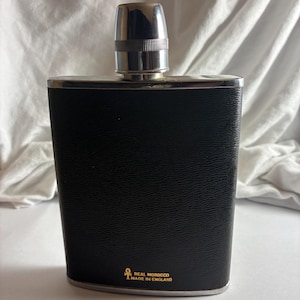 Op de afbeelding: Een zwartleren heupfles met een zilverkleurige dop en afwerking. De fles heeft een rechthoekige vorm met afgeronde randen. De tekst "REAL MOROCCO MADE IN ENGLAND" is in goud aan de onderkant gedrukt.