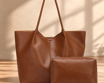 May include: A smooth, medium-brown leather tote bag with two long shoulder straps and a central vertical seam detail, accompanied by a matching rectangular zippered pouch positioned in front of the larger bag.