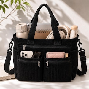 Può includere: Una borsa tote in velluto a coste nero con due manici superiori e una tracolla rimovibile. L'esterno presenta due tasche frontali con cerniera contenenti uno smartphone, occhiali da sole e un portafoglio. Lo scomparto principale contiene una borraccia, un taccuino e delle cuffie.