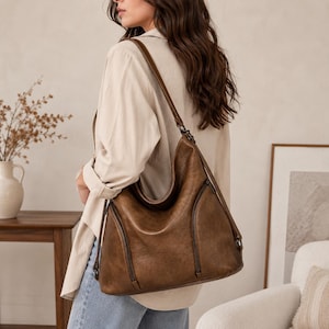Op de afbeelding: Een bruine hobo-schoudertas met een soepel silhouet, een enkele brede schouderband en twee verticale ritsvakken aan de voorzijde. De tas is uitgevoerd met een gladde, matte afwerking en is voorzien van donkerkleurige metalen hardware.