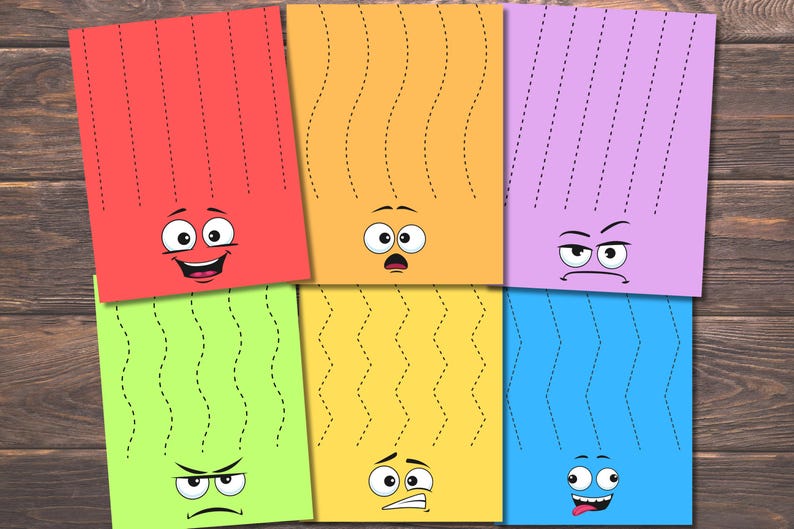 May include: A collection of colorful, cartoon-faced paper sheets with dotted lines for cutting practice. The sheets are red, orange, purple, green, yellow, and blue, each with a different facial expression. The background is a wooden surface.