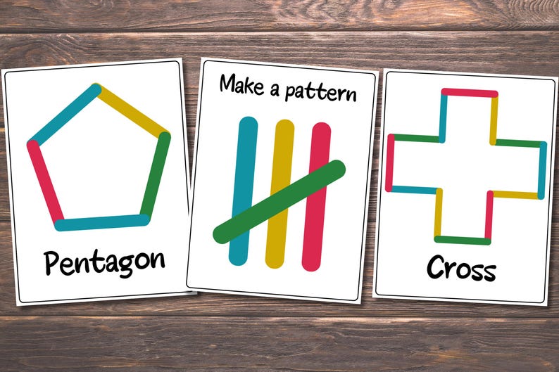 May include: Three white cards with colorful popsicle stick designs. One card shows a pentagon, another says "Make a pattern" with vertical lines, and the last shows a cross. The cards are on a wooden surface.