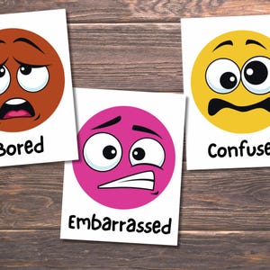 May include: Three cards with cartoon faces expressing emotions. One card shows a brown face labeled "Bored", a pink face labeled "Embarrassed", and a yellow face labeled "Confused". The cards are on a wooden surface.