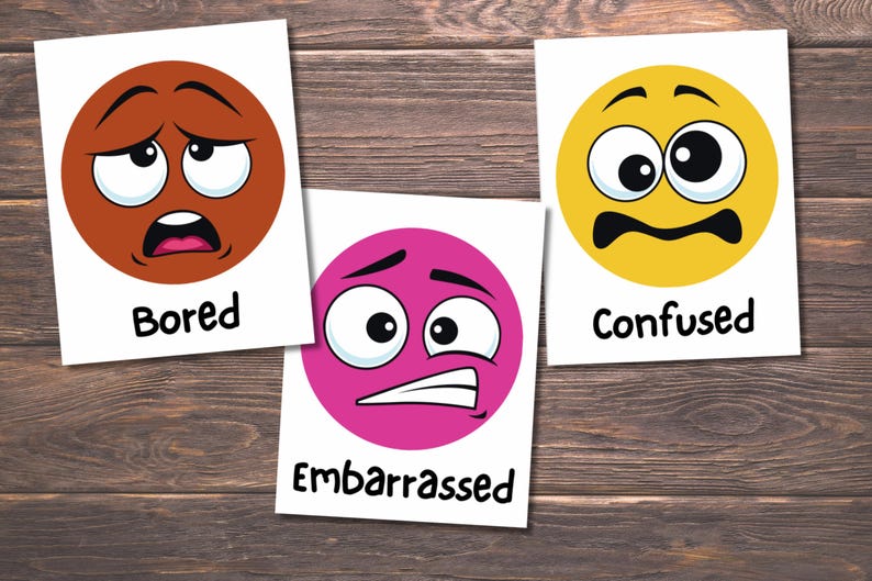 May include: Three cards with cartoon faces expressing emotions. One card shows a brown face labeled "Bored", a pink face labeled "Embarrassed", and a yellow face labeled "Confused". The cards are on a wooden surface.