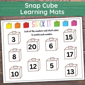 May include: A white learning mat with the text "Snap Cube Learning Mats" and "STACK IT". The mat features numbered cubes and instructions to match the numbers with stack cubes. The background is a wooden surface.
