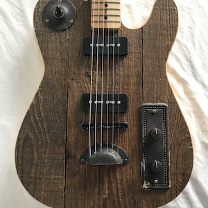 May include: A rustic electric guitar with a wooden body. The guitar has a weathered, brown wood grain finish, black pickups, and metal hardware. The neck is light-coloured wood with frets and strings.