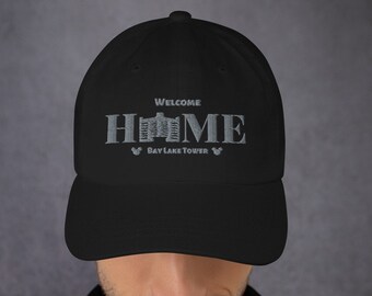 Könnte beinhalten: Schwarze Baseballkappe mit einem grauen gestickten Design, das "Willkommen Zuhause" mit einem stilisierten Bild des Bay Lake Towers und Mickey Mouse-Ohren auf beiden Seiten sagt.