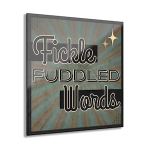 May include: A framed art print with the words "Fickle Fuddled Words" in a vintage style. The background has a teal and orange burst design with two gold star accents. The frame is black.