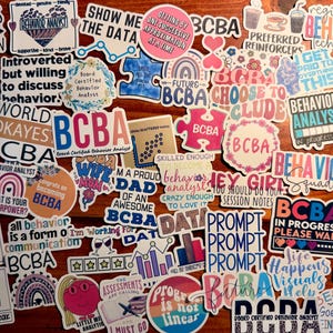 May include: A collection of colorful stickers with various designs and text related to behavior analysis. The stickers feature phrases like "BCBA," "Behavior Analyst," and motivational quotes. The stickers are on a wooden surface.