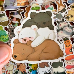 May include: A die-cut sticker featuring a stack of three cartoon bears. A dark brown bear lies at the bottom, a white bear rests in the middle, and a smaller dark gray bear sleeps on top. The bears have closed eyes and a soft, rounded appearance.