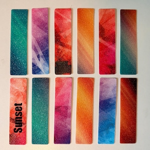 May include: Twelve rectangular bookmarks arranged in two rows, each featuring a unique abstract gradient pattern with vibrant colors including teal, purple, red, orange, and blue. One bookmark in the bottom left corner has the word Sunset printed.