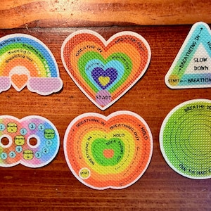 May include: A collection of colorful, embroidered patches with text related to breathing exercises. The patches are in various shapes, including a rainbow, hearts, a triangle, and circles. The text includes phrases like "Breathe In," "Breathe Out," and "Slow Down."