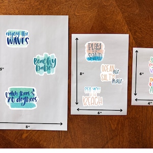 May include: Three sheets of stickers with beach-themed phrases. The largest sheet is 11.5 inches long and 8 inches wide, with stickers that say "enjoy the WAVES", "Beachy Babe", and "Palm trees & 80 degrees". The second sheet is 8 inches long and 6 inches wide, with stickers that say "PLAY in the SAND", "SEA YOU at the BEACH", and "OCEAN air SALTY hair". The smallest sheet is 6 inches long and 4 inches wide, with stickers that say "YOUR feelings are VALID" and "You Are NOT ALONE".