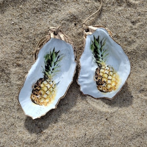 Puede incluir: Dos conchas de ostra blancas con ribetes dorados, cada una con un diseño de piña pintado. Las piñas son amarillas con hojas verdes y están sobre un fondo blanco.