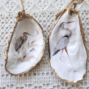 May include: Two oyster shell ornaments with painted heron designs. The shells have a gold-painted edge and are suspended by twine. The heron illustrations are in shades of grey and brown, set against a white background.