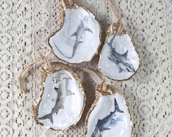 May include: Four oyster shell ornaments featuring detailed shark illustrations. Each shell has a white interior, a gold-painted rim, and a natural jute twine loop attached at the top for hanging. The sharks are depicted in various swimming poses.