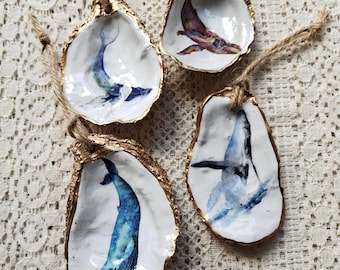 May include: Four oyster shell ornaments featuring hand-painted whale illustrations in shades of blue, purple, and brown. Each shell has a gold-painted edge and a natural jute twine loop attached at the top for hanging, showcasing detailed marine art.