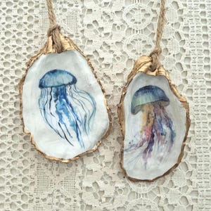 May include: Two oyster shells with gold-painted edges, each featuring a watercolor jellyfish design. One jellyfish is blue, the other is a mix of colors. Both shells have a natural rope for hanging.