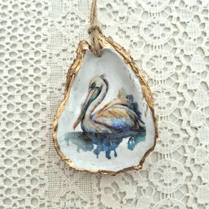 May include: An oyster shell ornament featuring a watercolor painting of a pelican. The pelican is depicted with brown, white, and blue feathers, resting on a blue water surface. The shell has a gold-painted edge and a natural rope for hanging.