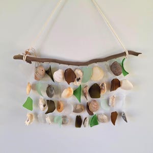 May include: A handcrafted wall hanging featuring a natural driftwood branch. Strands of seashells and sea glass in shades of green, brown, and white are suspended from the branch, creating a coastal-themed decorative piece.