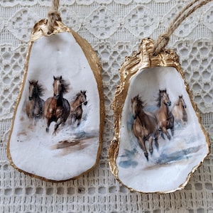以下が含まれることがあります： 金で塗られた2つのカキの殻に、手描きで描かれた走る馬の絵が描かれています。馬は茶色と白で、白い背景に描かれています。