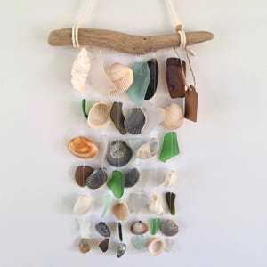 May include: A coastal-themed wall hanging featuring a driftwood base with various seashells and sea glass pieces suspended by strings. The arrangement includes a variety of colors and shapes, creating a natural and organic aesthetic.