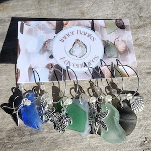 May include: A set of six wine charms with sea-themed pendants. Each charm features a piece of sea glass in various colors, including blue and green, and a silver-toned charm, such as a starfish or seashell. The card behind the charms reads "Inner Banks Adventures."