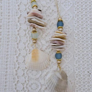 May include: Two seashell ornaments hanging from beige string. Each ornament features a stack of various seashells, gold beads, and a large shell at the bottom. One has a blue bead, the other a light green bead.
